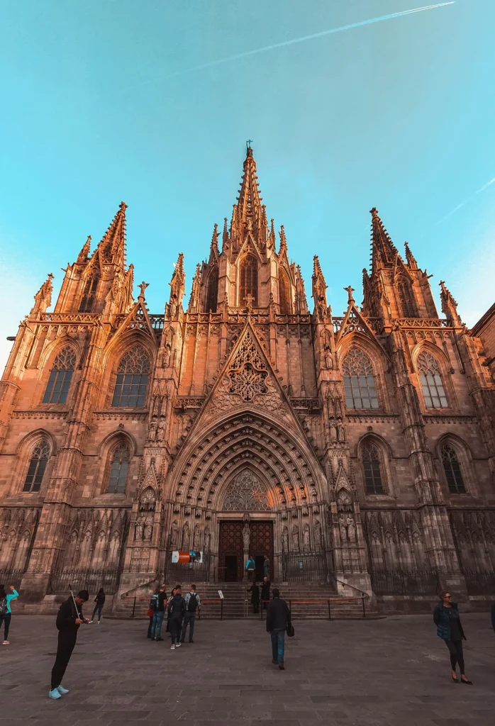People stand in front of a large Gothic cathedral with pointed arches, intricate carvings, and tall spires under a clear sky, while nearby the modern L’Auditori Barcelona adds contrast to the historic scene.